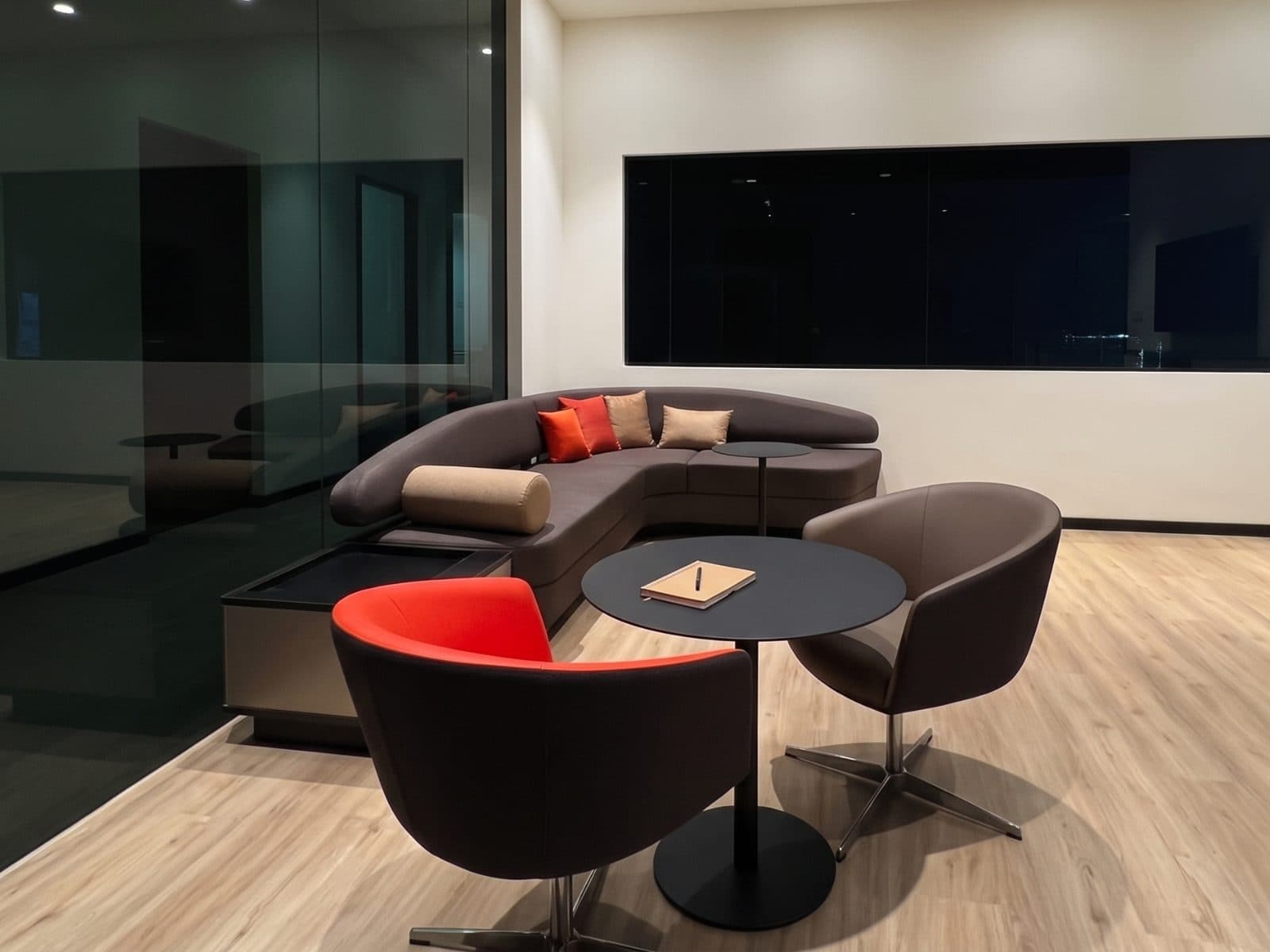A lounge with a curved sofa, low round tables and tub chairs in red and brown, set beside a glazed partition.